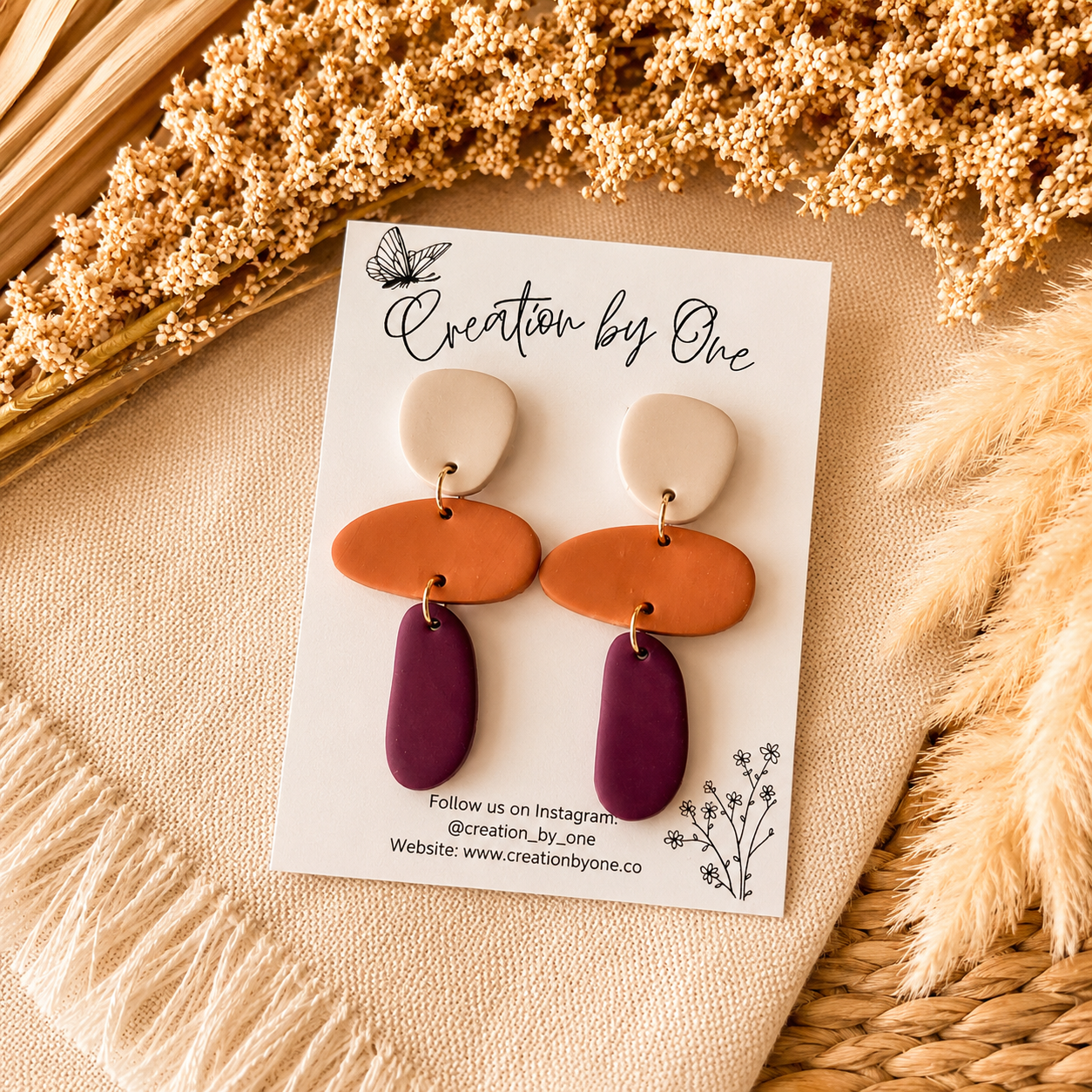 Pair of earrings on a 'Creation by One' branded card with dried flowers in the background.