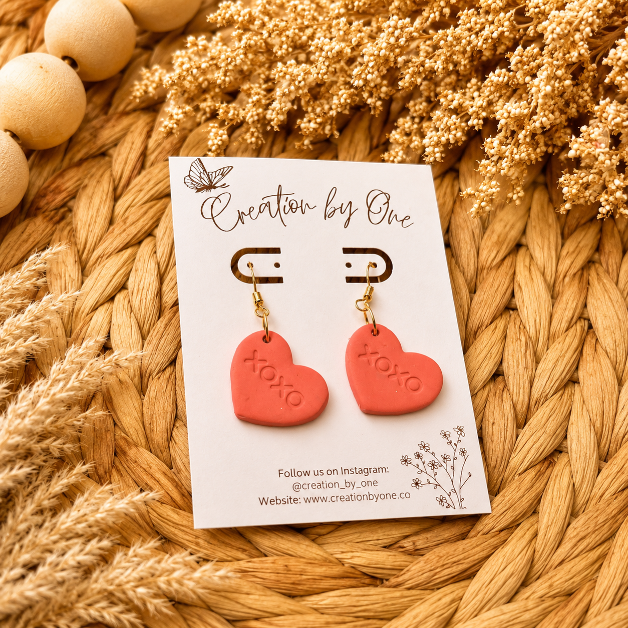 Red heart-shaped earrings on a 'Creation by One' branded card with dried flowers and beads in the background.