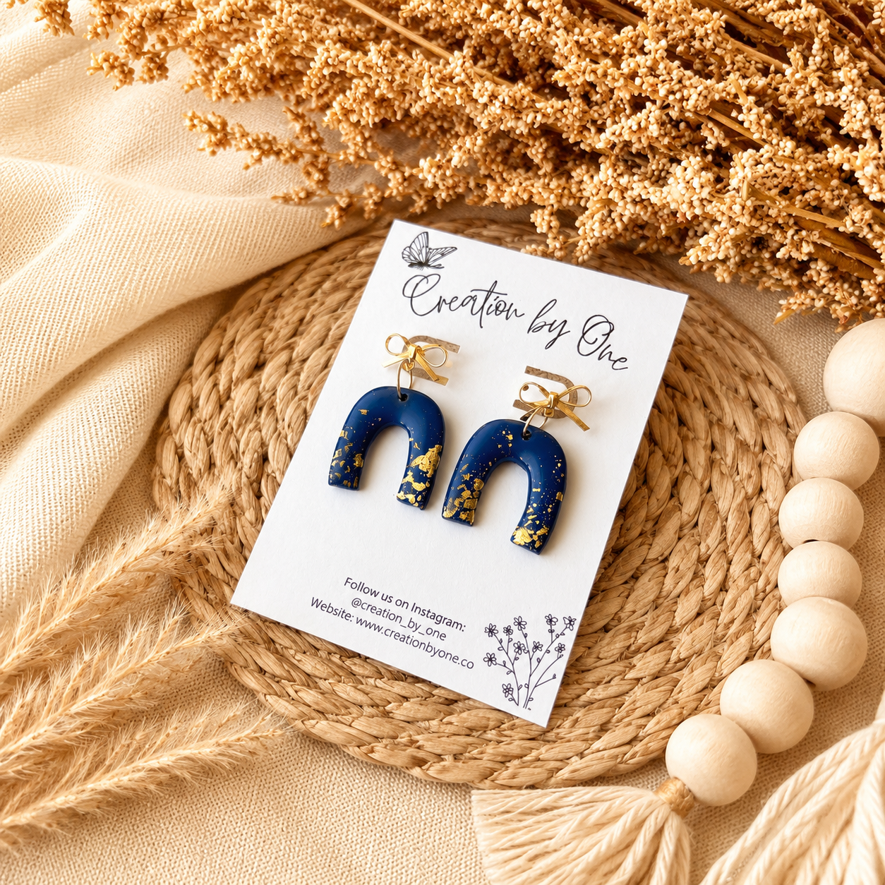 Blue and gold earrings on a 'Creation by One' branded card with dried flowers and beads in the background.