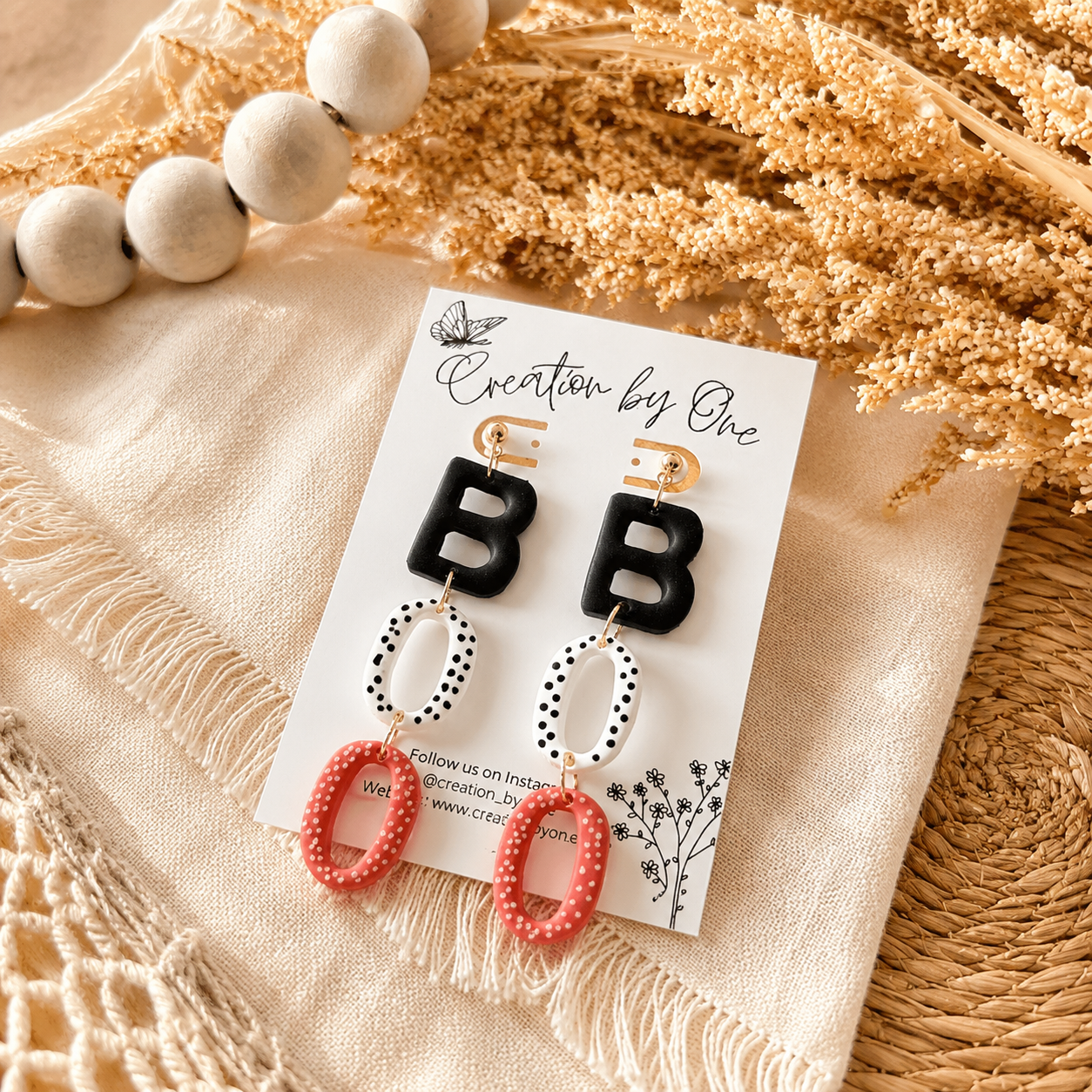 Pair of earrings with 'BB' design on a branded card, placed on a textured surface with dried flowers.