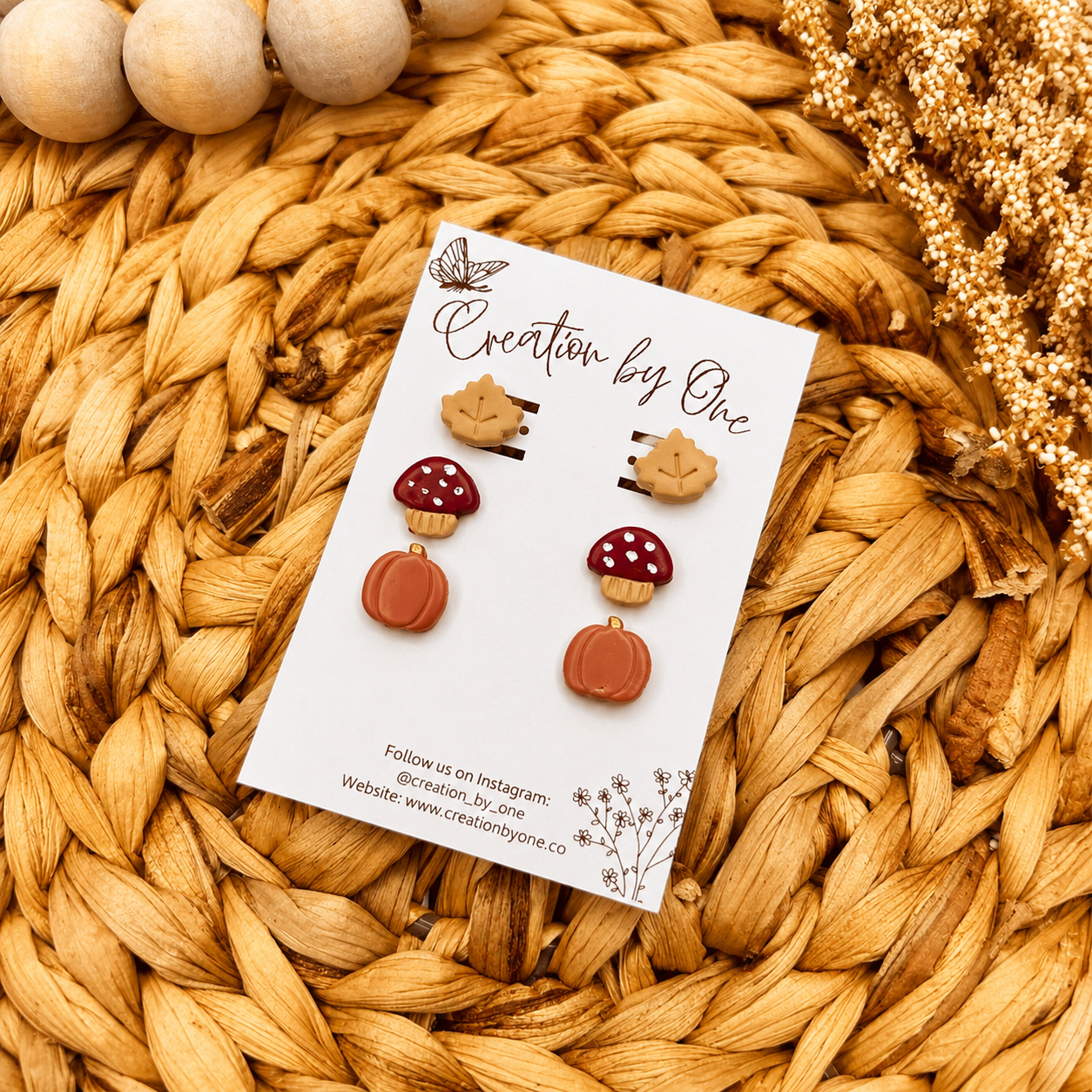 Handmade fall clay stud earrings by Creation by One featuring beige maple leaves, burgundy mushrooms, and dusty pumpkin studs displayed on a branded earring card with a neutral boho woven background, wooden beads, and dried florals.