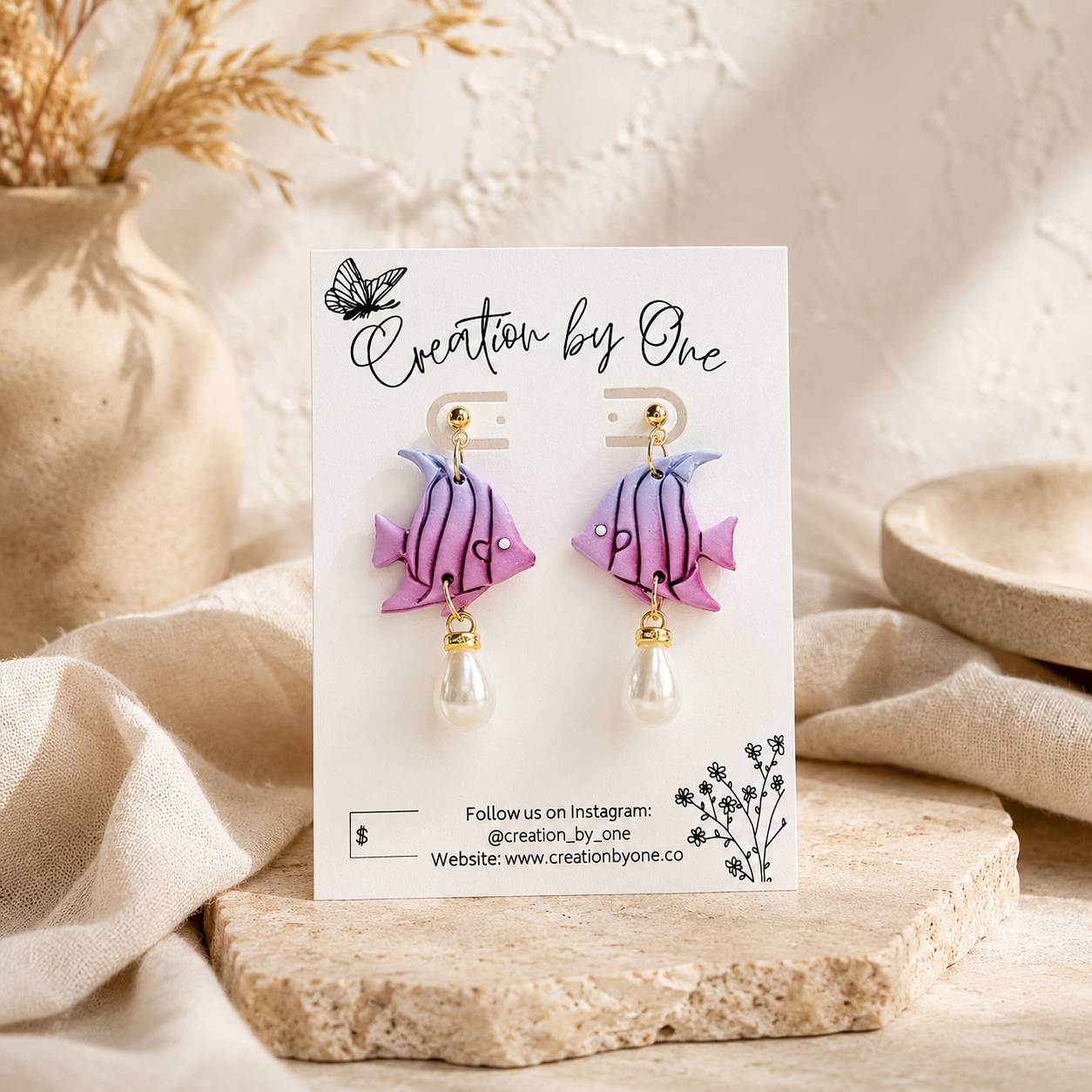A pair of dangling fish-shaped earrings displayed on a white card. The earrings have purple and pink striped fish bodies with small gold hooks and white pearl drops.