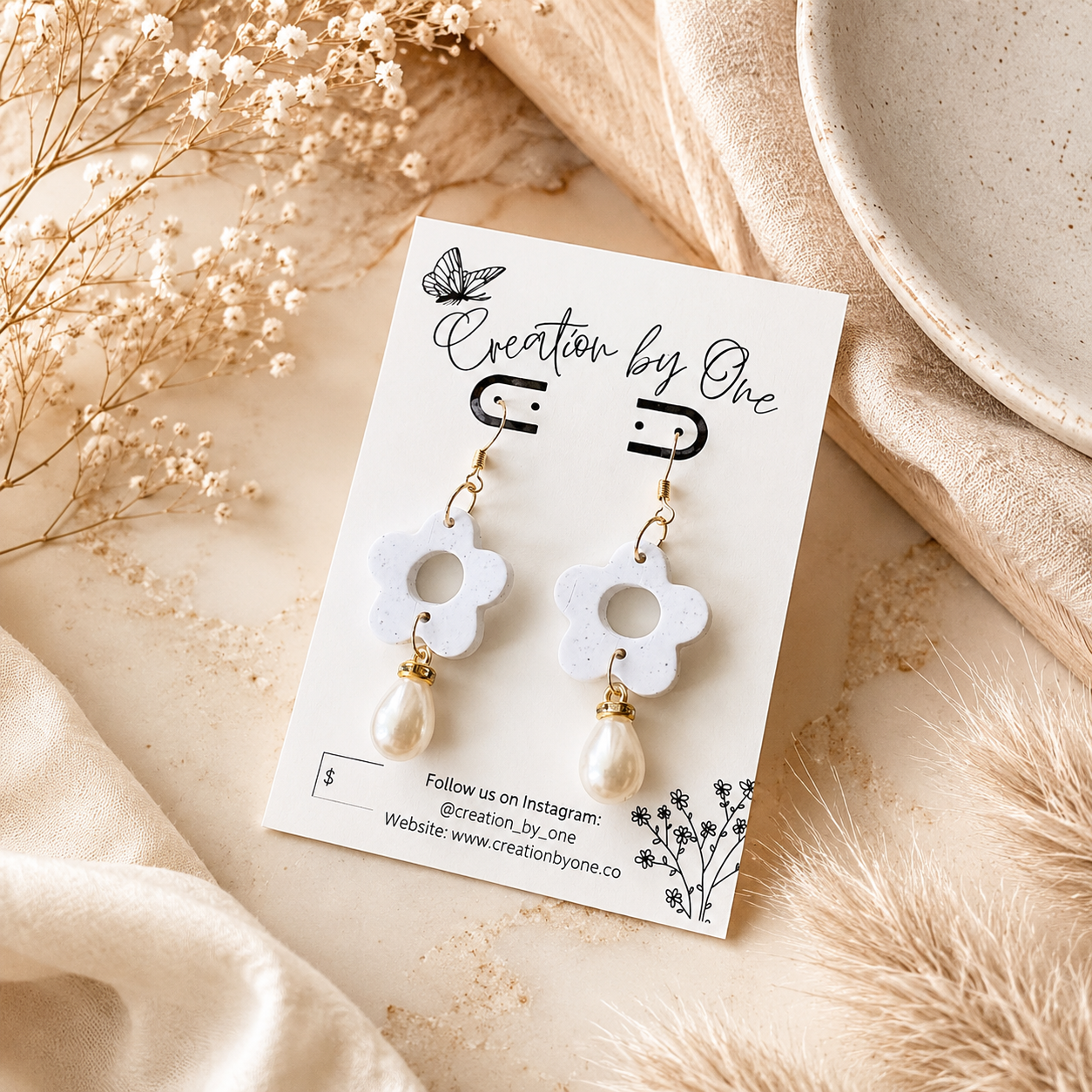 A pair of white flower-shaped drop earrings with gold-tone hardware and pearl-like dangling beads, displayed on a branded card over a soft beige fabric and dried floral arrangement.