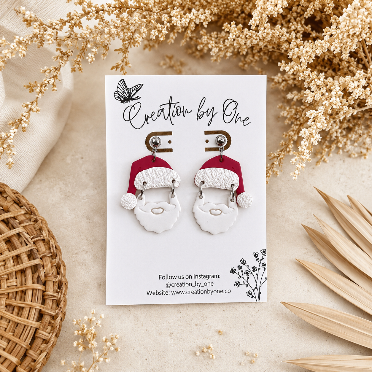 Handmade Santa Claus polymer clay dangle earrings on a Creation by One display card with a neutral boho background featuring dried florals and soft beige textures, perfect for Christmas outfits, holiday gifting, and festive winter accessories.