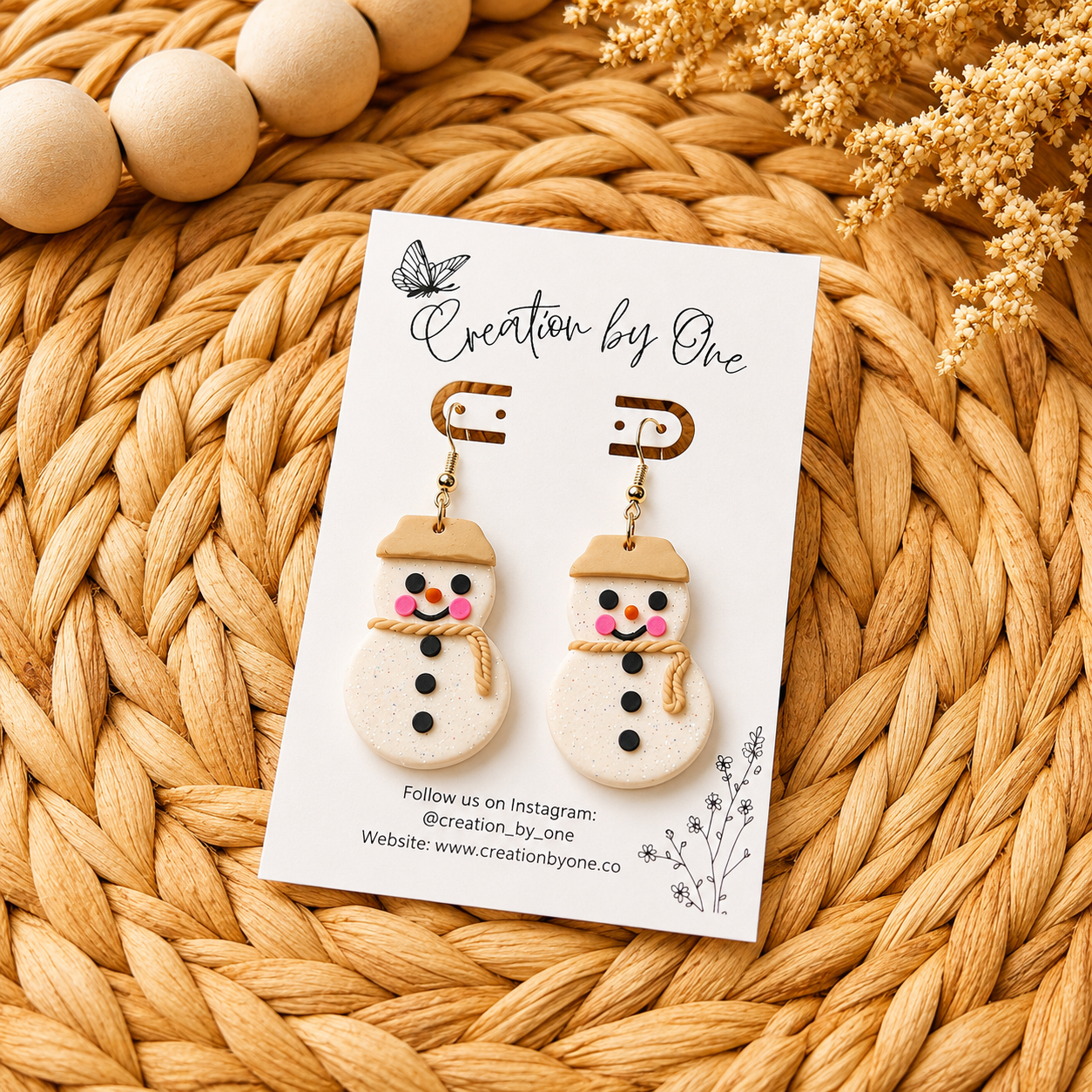 Handmade polymer clay snowman dangle earrings displayed on a branded jewelry card over a woven boho straw background with natural wooden beads and dried flowers. Cute Christmas earrings with gold hooks, rosy cheeks, and a rustic scarf design.