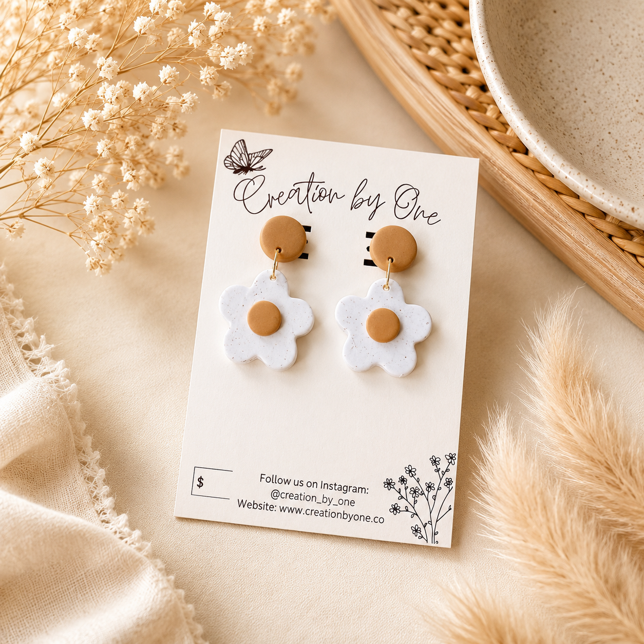 A pair of handmade daisy earrings with white felt petals and tan centers, displayed on a white card labeled 'Creation by One' on a beige fabric surface with dried flowers and a woven tray.