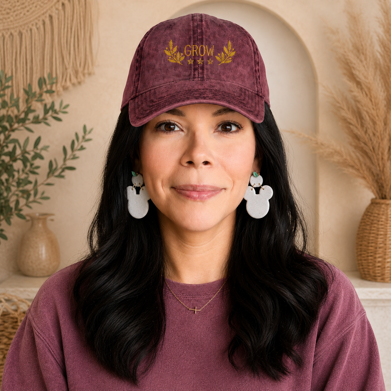 A woman with long dark hair wearing a dusty burgundy baseball cap with gold embroidered text reading 'GROW WELL' and white Mickey-shaped earrings, posed in a warm, boho-style room setting.