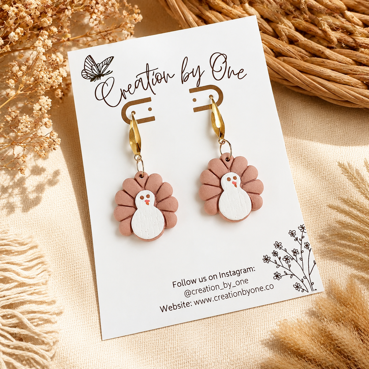 Pair of earrings on a 'Creation by One' branded card with dried plants in the background