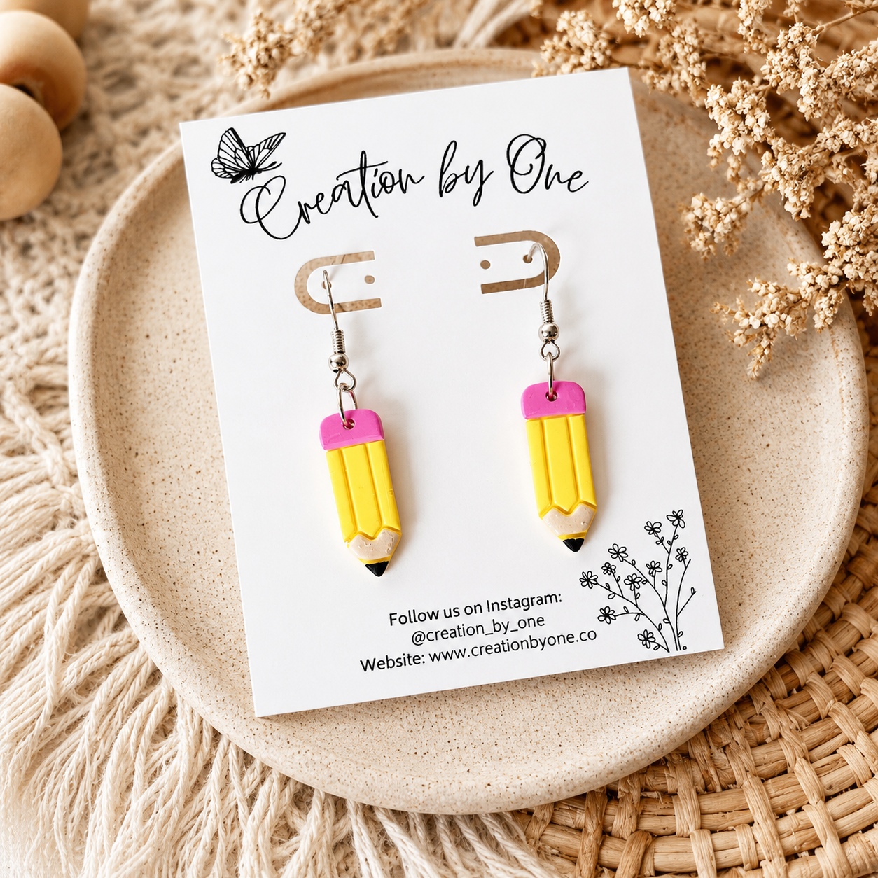 Yellow and pink pencil-shaped earrings on a 'Creation by One' branded card, placed on a textured surface with dried flowers.