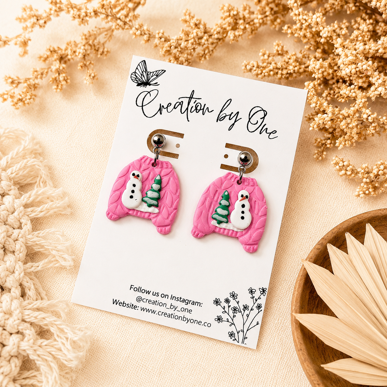 Handmade pink polymer clay dangle earrings shaped like cozy knit sweaters with snowman and Christmas tree details, displayed on a Creation by One jewelry card against a neutral boho linen background with dried florals.