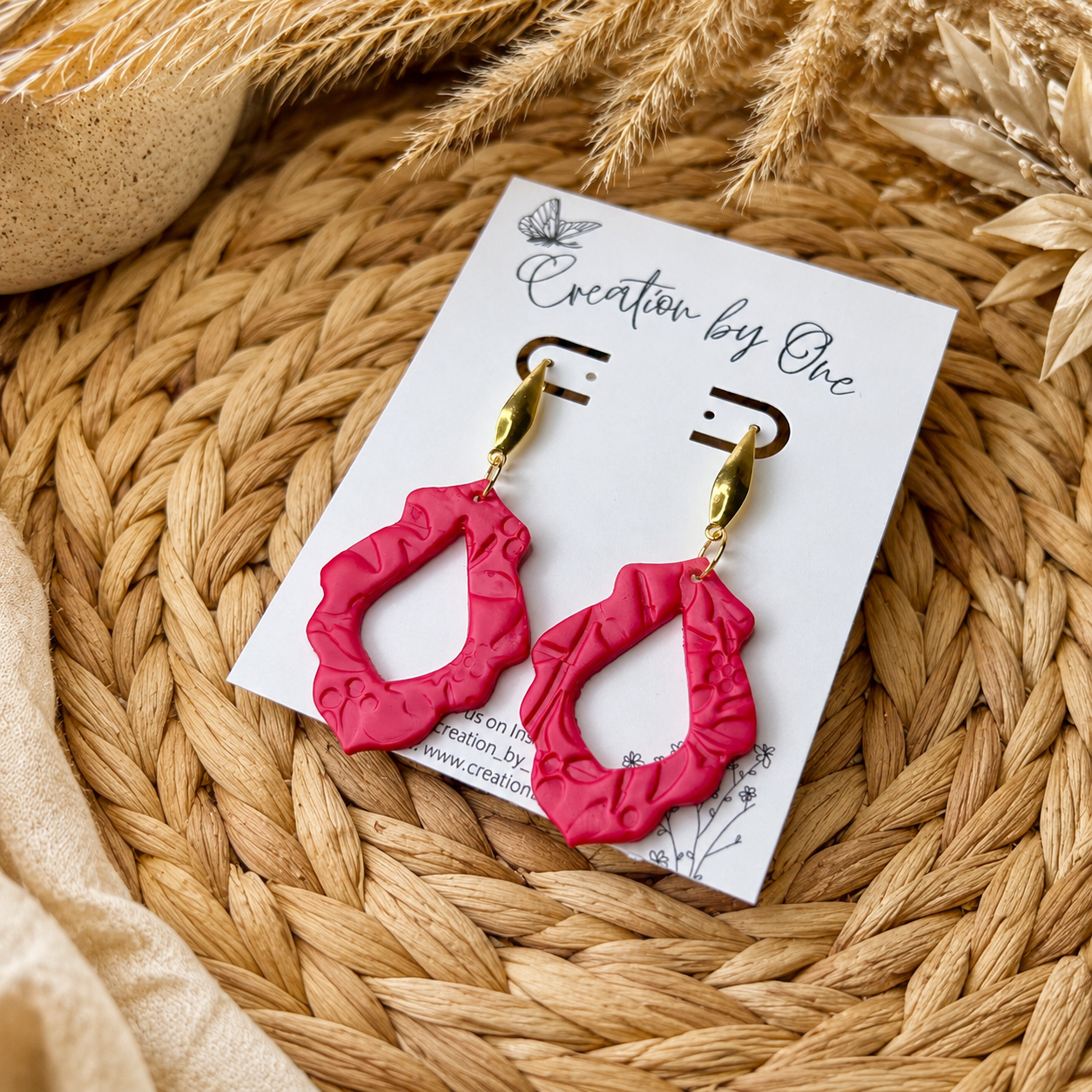 Pink earrings on a white card with 'Creation by One' branding, placed on a woven surface.