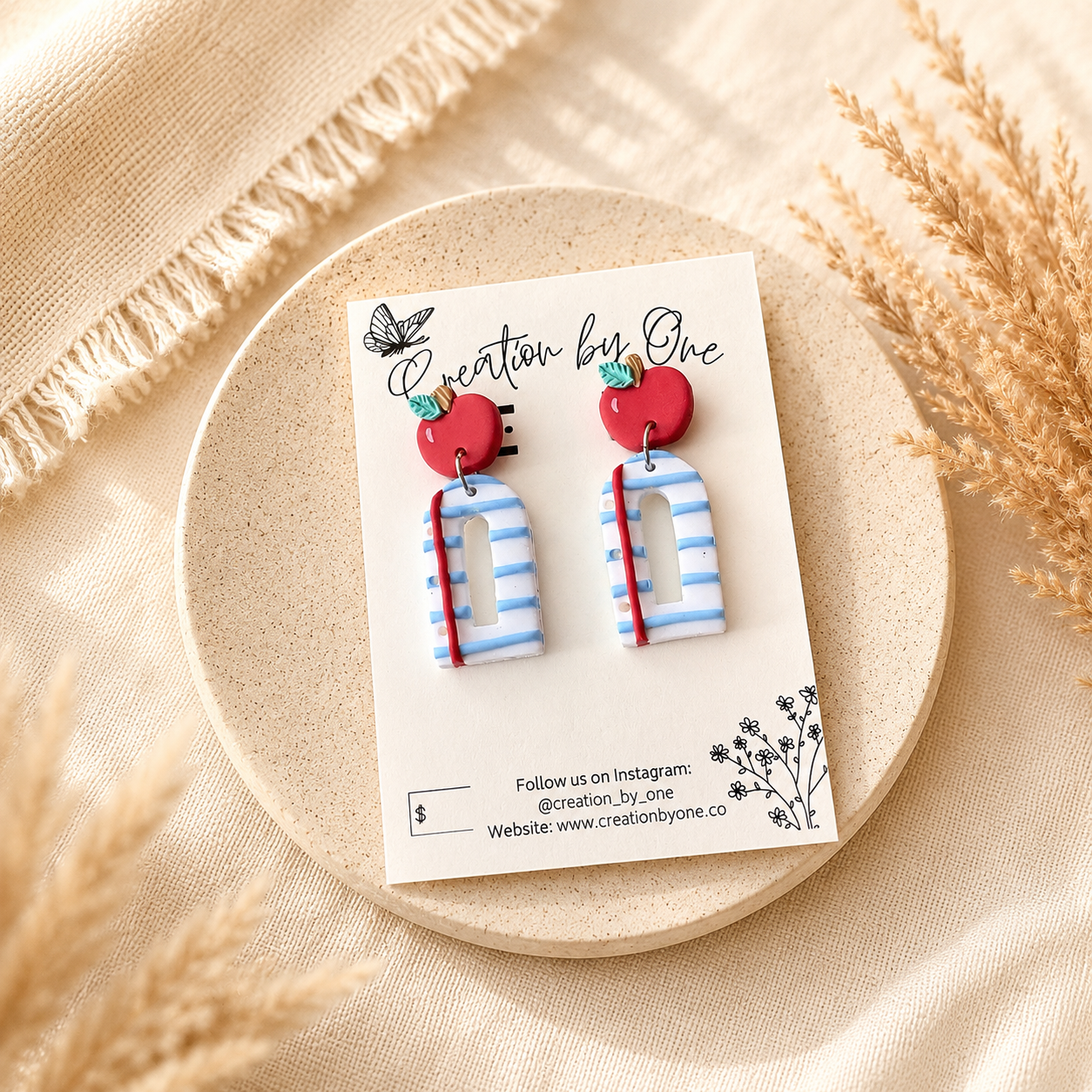 Handmade polymer clay teacher earrings displayed on a Creation by One jewelry card featuring bright red apple studs paired with blue-and-white striped notebook-style dangles accented with a red pencil detail. The card is styled on a soft neutral stone tray with warm beige linen fabric and dried pampas grass, creating a clean, modern boho product display with natural lighting.