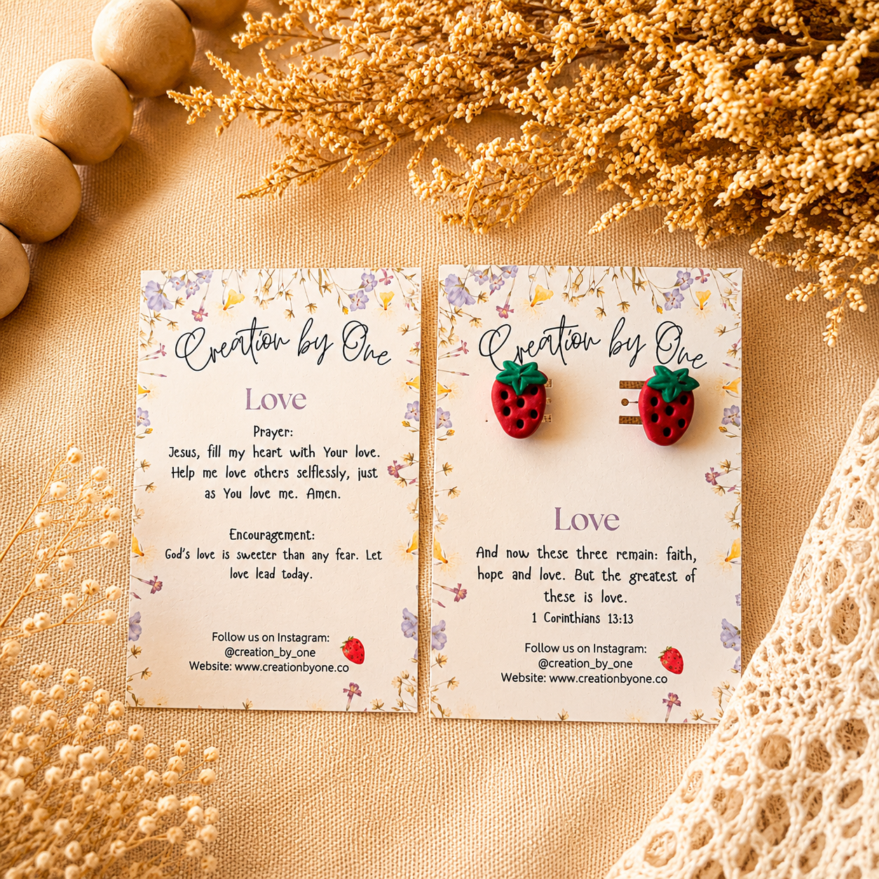 Two strawberry-shaped earrings on cards with text and branding, placed on a decorative surface with flowers and stones.