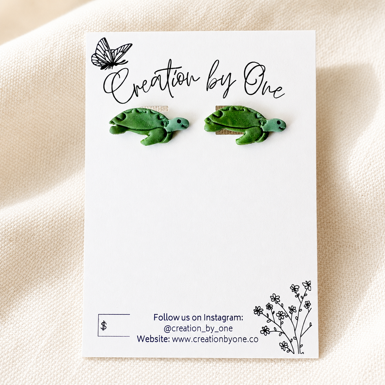 A pair of green turtle stud earrings displayed on a white card labeled 'Creation by One,' placed on a soft cream fabric background.