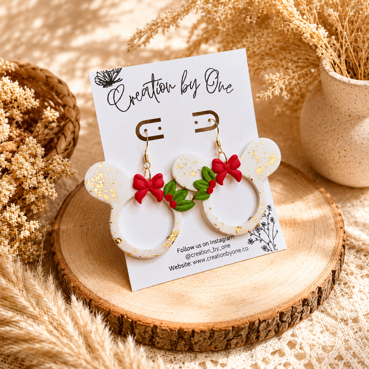 Handmade white Christmas mouse-shaped dangle earrings with gold leaf flakes, red bows, and holly accents displayed on a jewelry card against a soft neutral boho fabric background with dried florals.