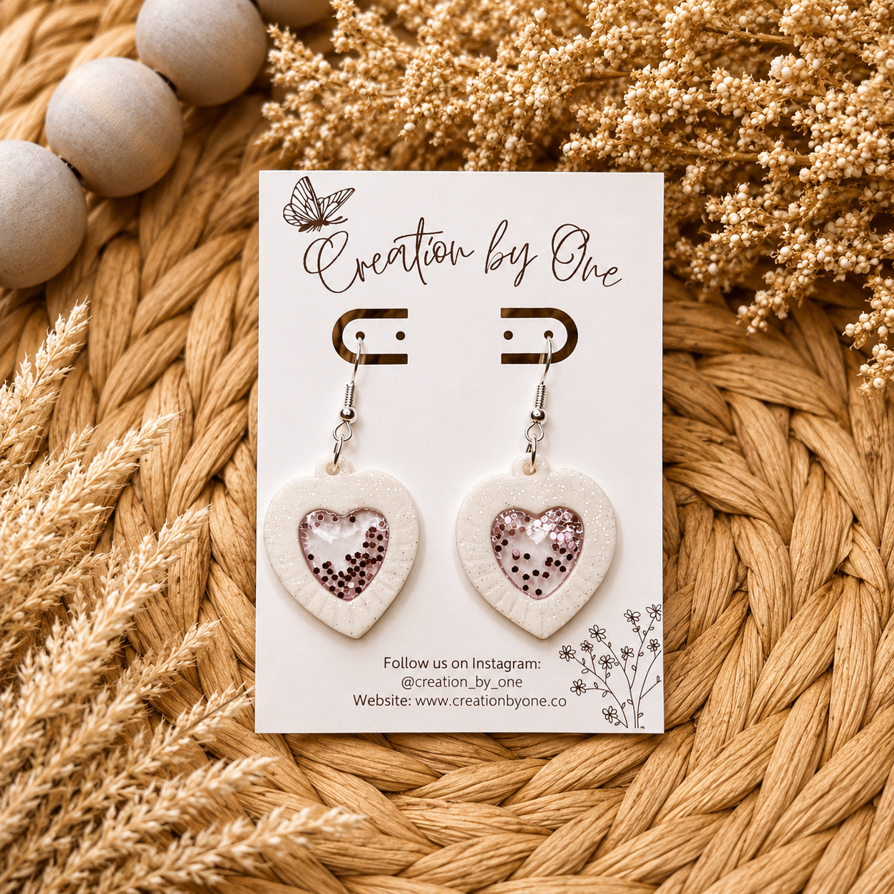 Heart-shaped earrings on a card with 'Creation by One' branding, placed on a woven surface with dried flowers.