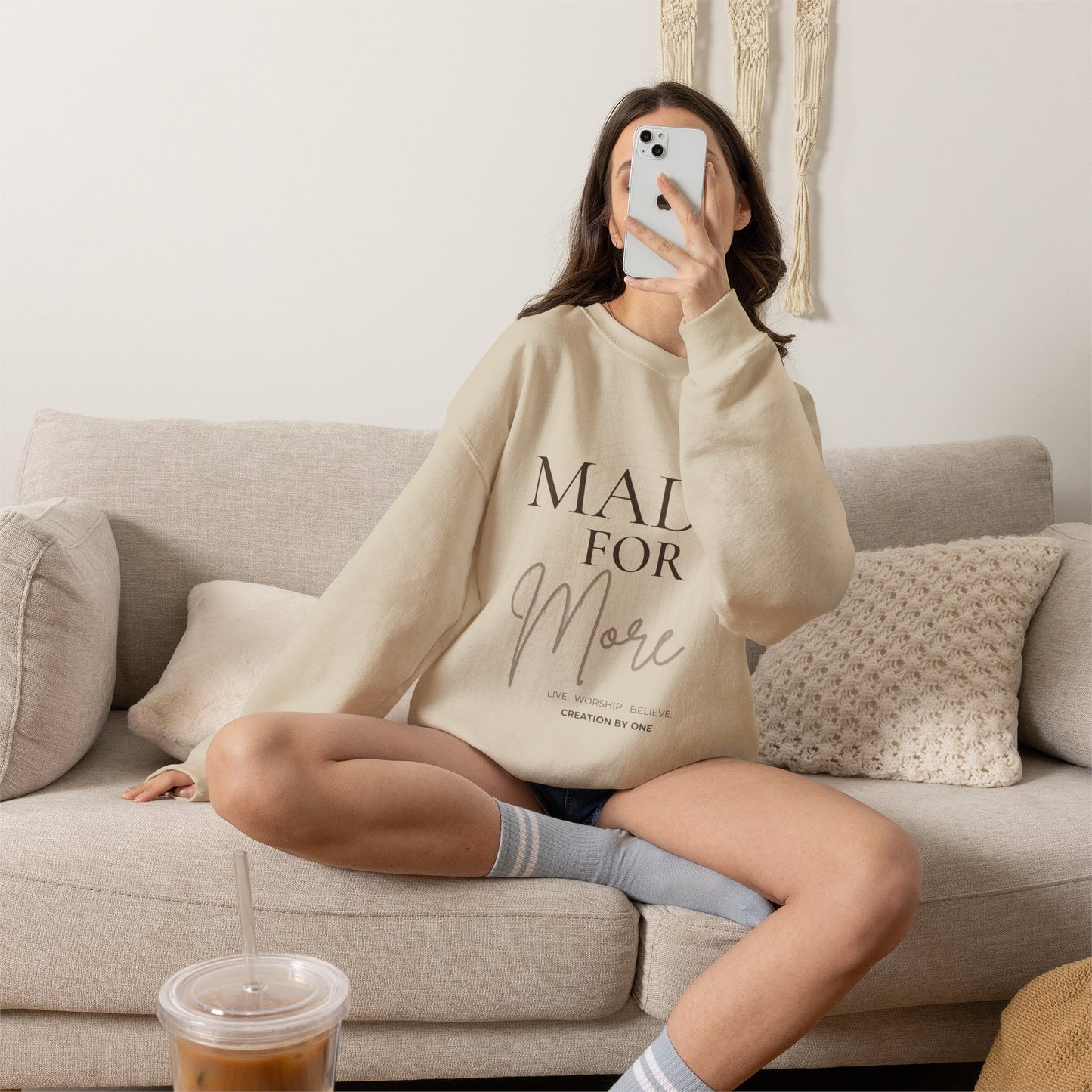 Person wearing a beige sweatshirt with text, sitting on a couch with a cup of coffee.