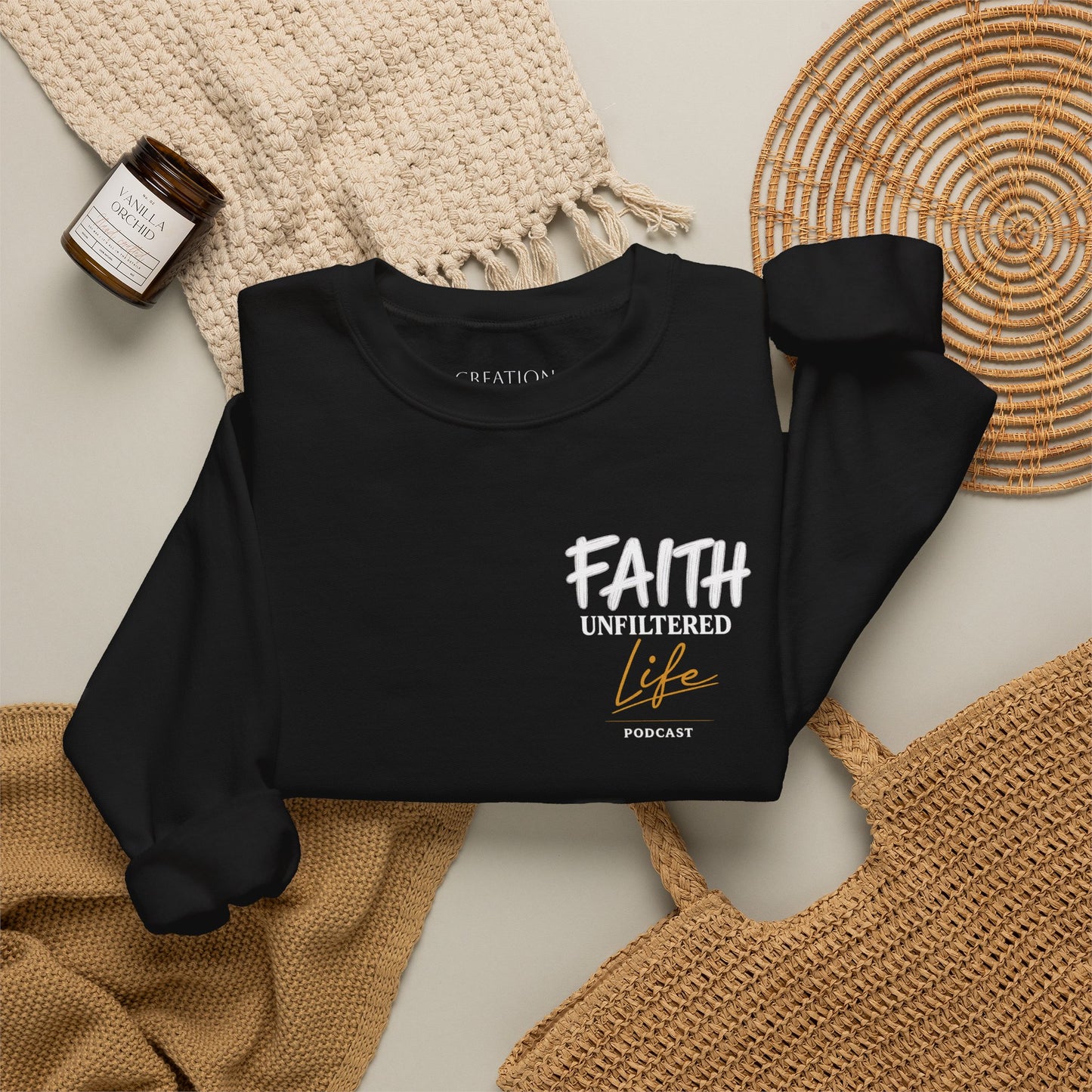 A black long-sleeve crewneck sweatshirt laid flat on a light beige surface, styled with a woven cream blanket, a small amber candle jar, and tan woven placemats. The sweatshirt has a front graphic reading “FAITH UNFILTERED Life PODCAST” in white and tan lettering.