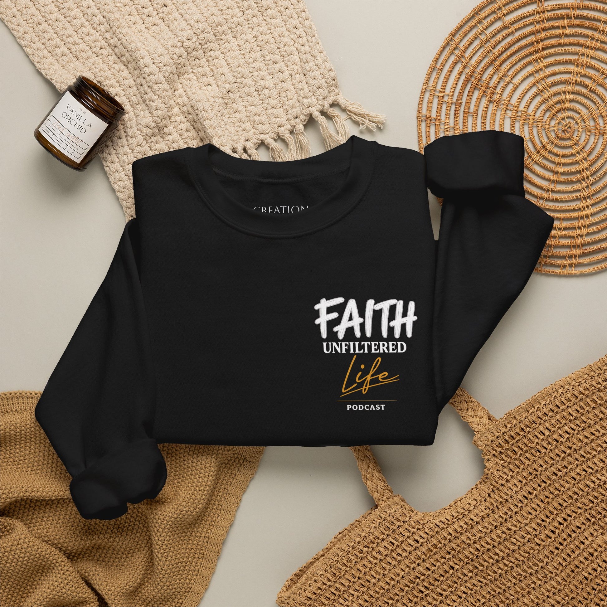 A black long-sleeve crewneck sweatshirt laid flat on a light beige surface, styled with a woven cream blanket, a small amber candle jar, and tan woven placemats. The sweatshirt has a front graphic reading “FAITH UNFILTERED Life PODCAST” in white and tan lettering.