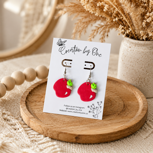 Red apple-shaped earrings on a card with 'Creation by One' branding, displayed on a wooden tray.