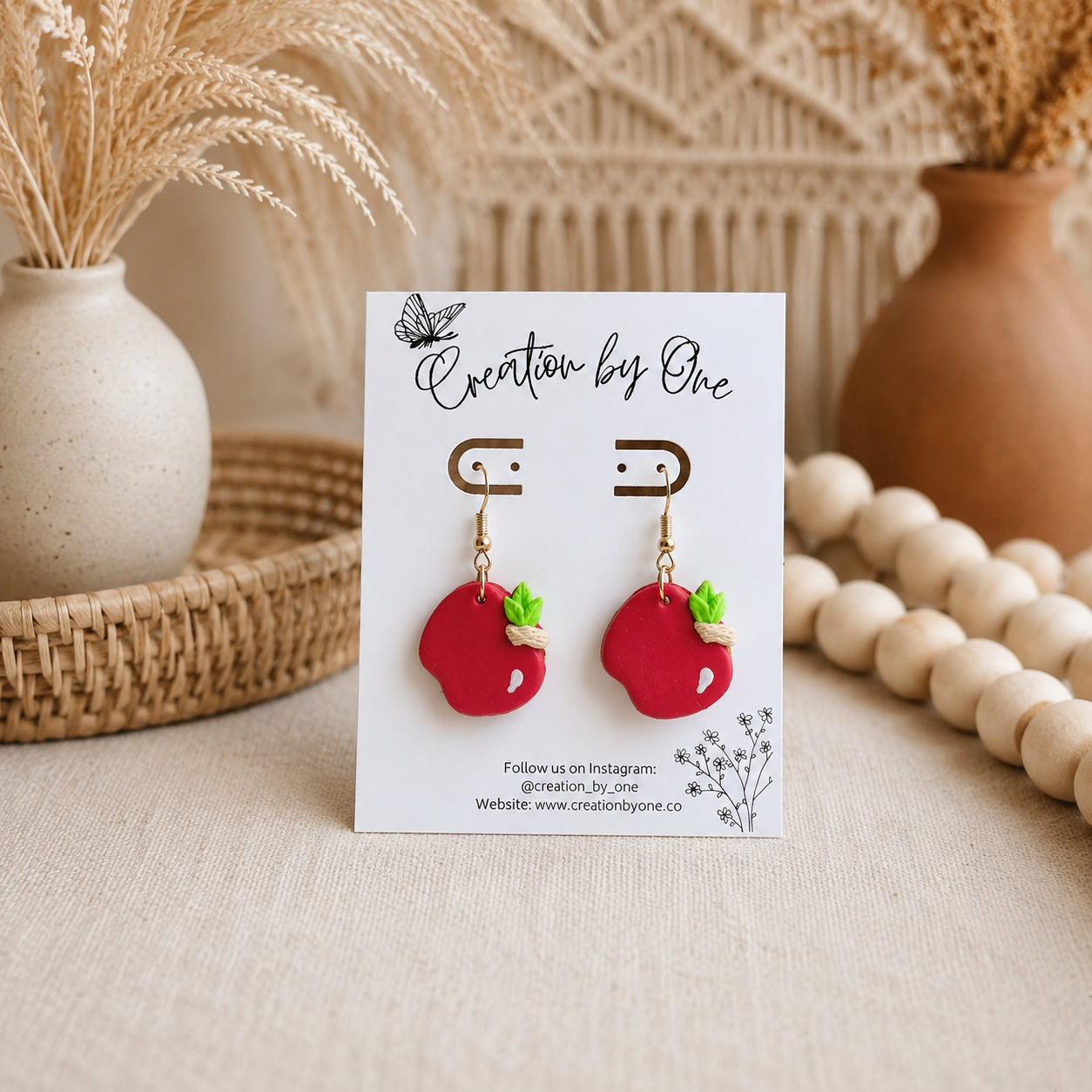 Red apple-shaped earrings on a card with 'Creation by One' branding, displayed on a neutral background.