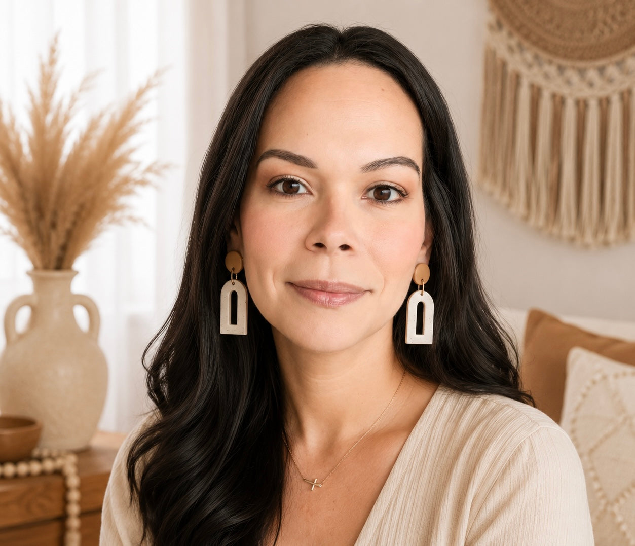 A woman with long dark hair wearing geometric white and brown drop earrings, posed in a warm, bohemian-style room with a vase of dried grass and a macrame wall hanging in the background.