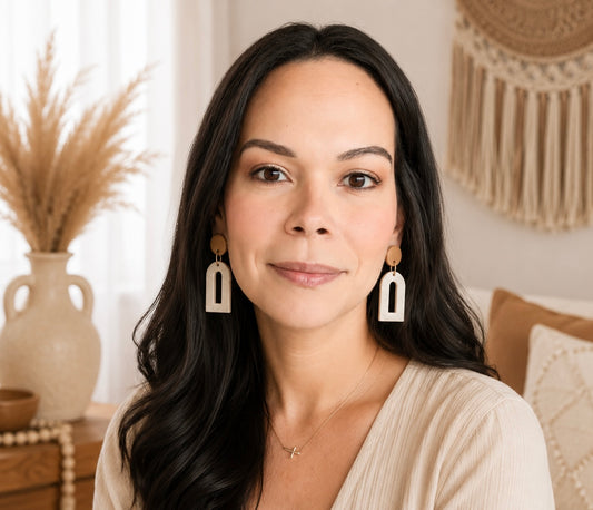 A woman with long dark hair wearing geometric white and brown drop earrings, posed in a warm, bohemian-style room with a vase of dried grass and a macrame wall hanging in the background.