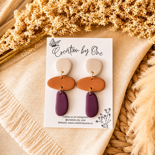 Pair of earrings on a 'Creation by One' branded card with dried flowers in the background.
