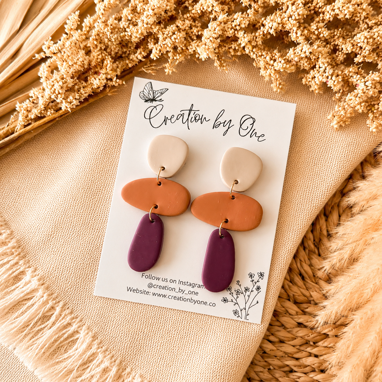 Pair of earrings on a 'Creation by One' branded card with dried flowers in the background.