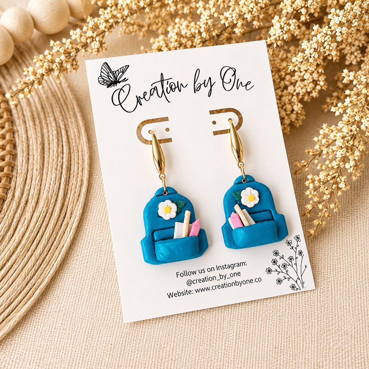 Blue backpack-shaped earrings on a 'Creation by One' branded card with floral decorations.
