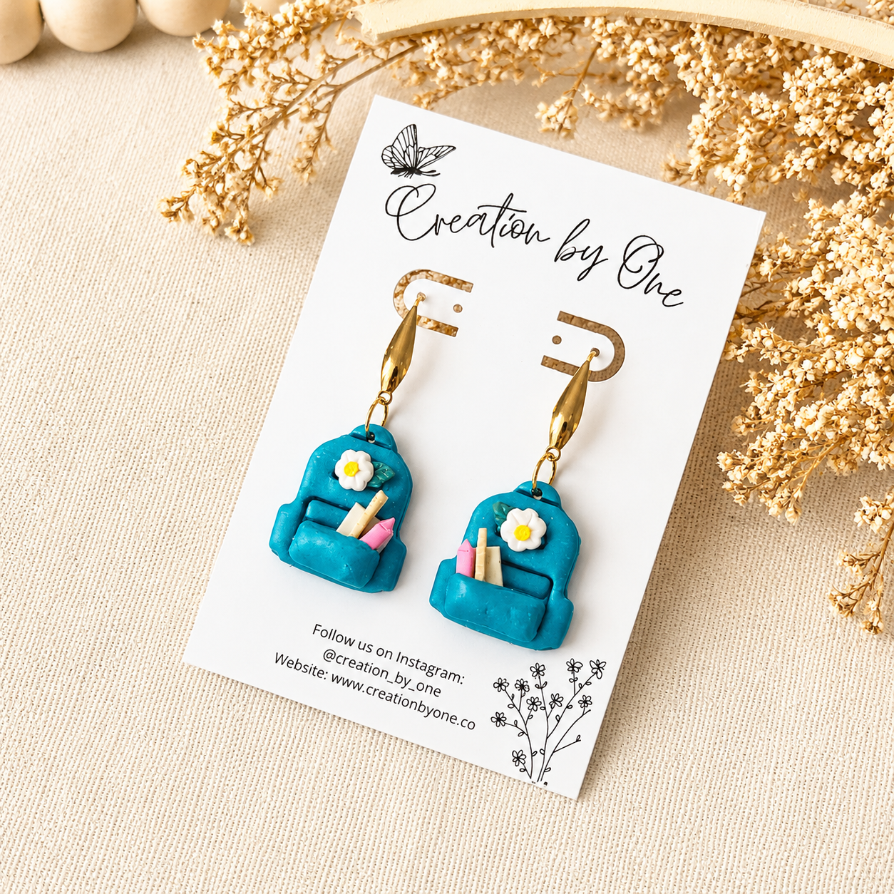 Turquoise earrings with gold accents on a 'Creation by One' branded card, surrounded by dried flowers.