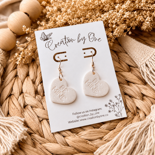 Heart-shaped earrings on a 'Creation by One' branded card with decorative elements.