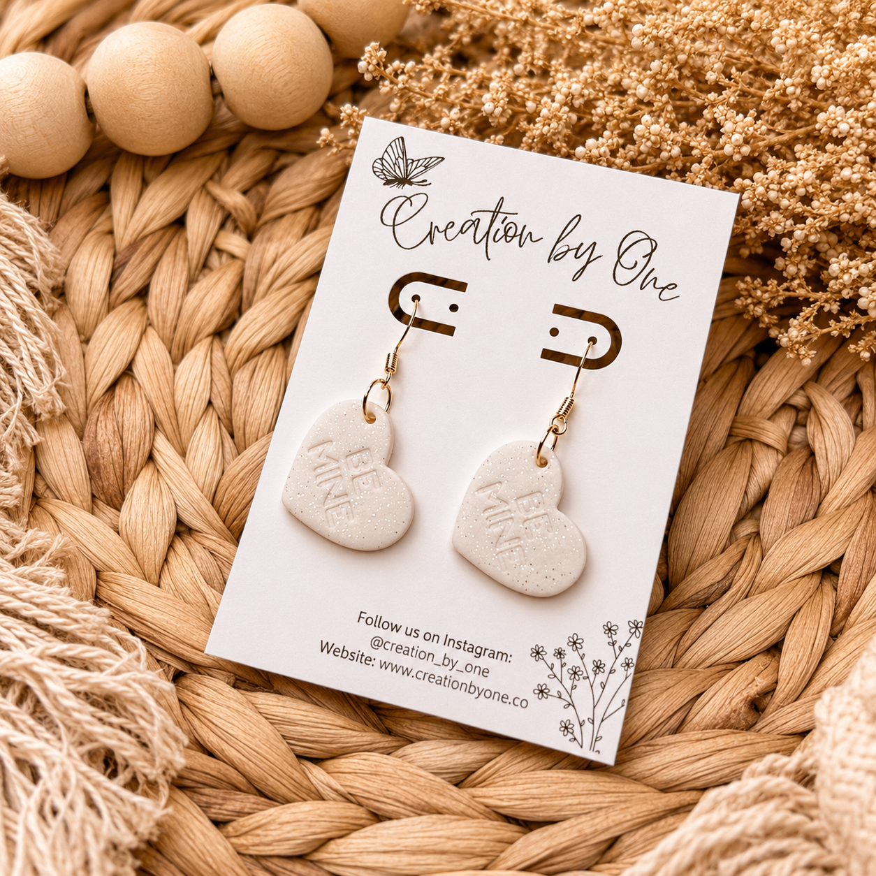 Heart-shaped earrings on a 'Creation by One' branded card with wooden beads and flowers in the background.