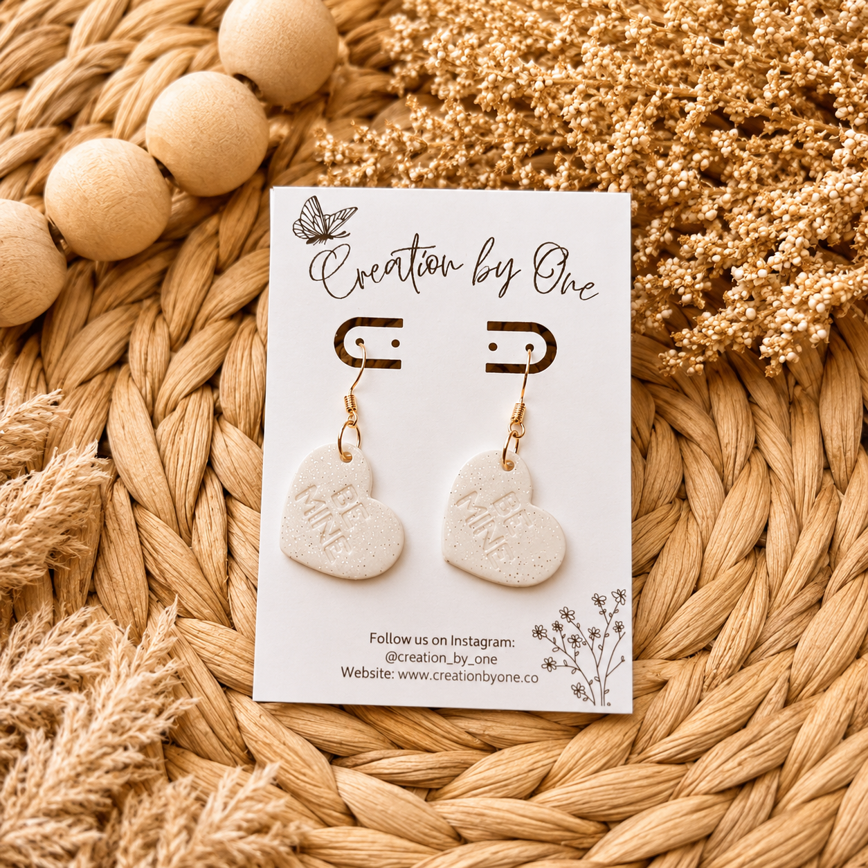 Heart-shaped earrings on a 'Creation by One' branded card with beige beads and dried flowers in the background.
