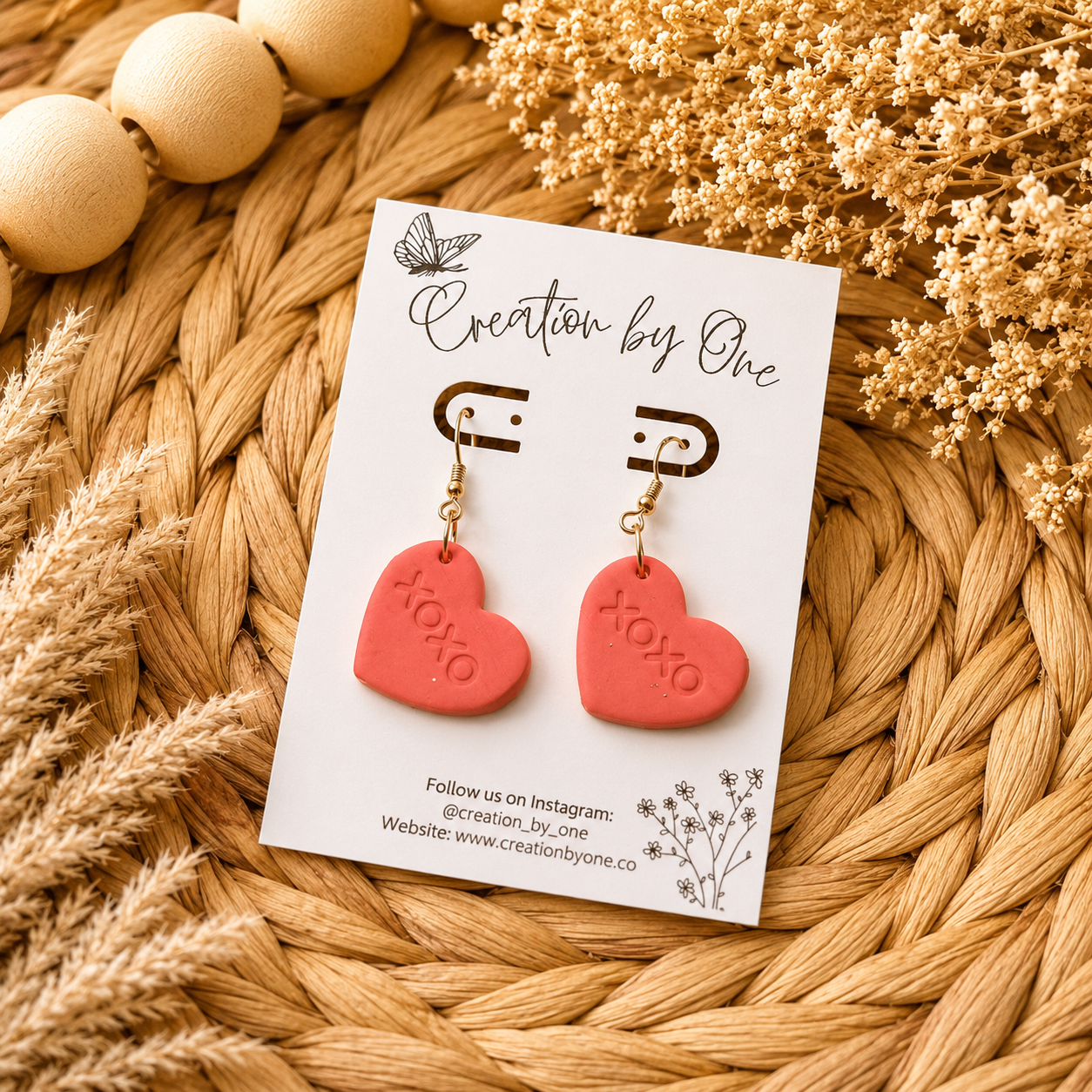 Red heart-shaped earrings on a 'Creation by One' branded card with dried flowers and beads in the background.