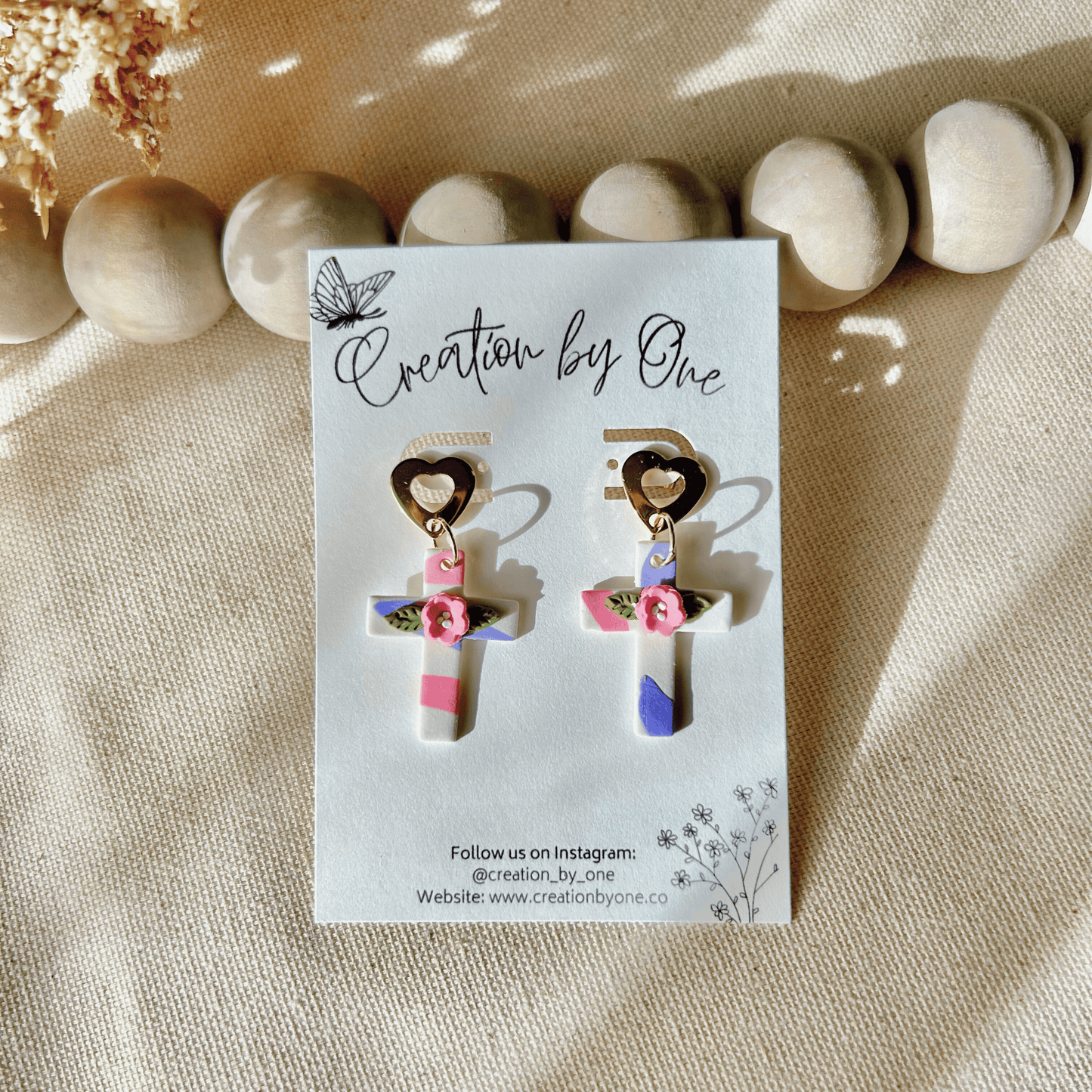 A pair of handcrafted polymer clay earrings with a cross design, featuring 14K gold-plated heart-shaped studs in white, pink, and purple colors, displayed on a beige fabric surface.