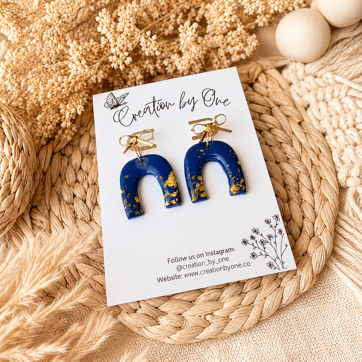 Blue earrings with gold accents on a 'Creation by One' branded card, placed on a woven mat with dried flowers.