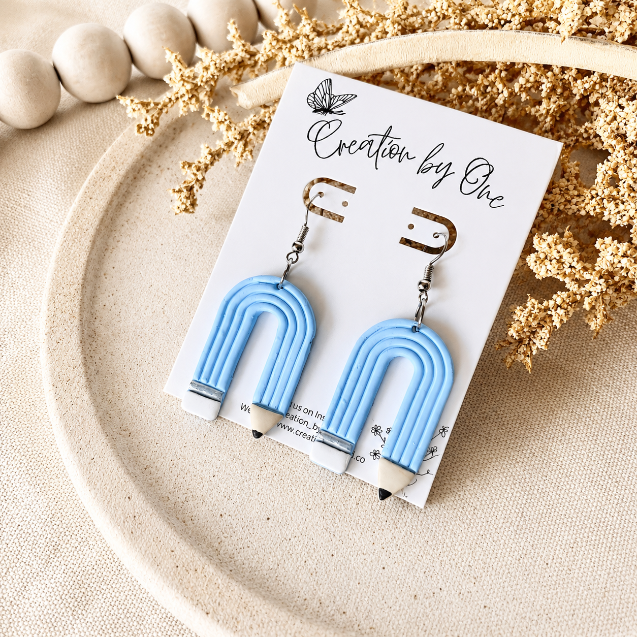 Blue U-shaped earrings on a card with 'Creation by One' branding, placed on a textured surface with decorative elements.