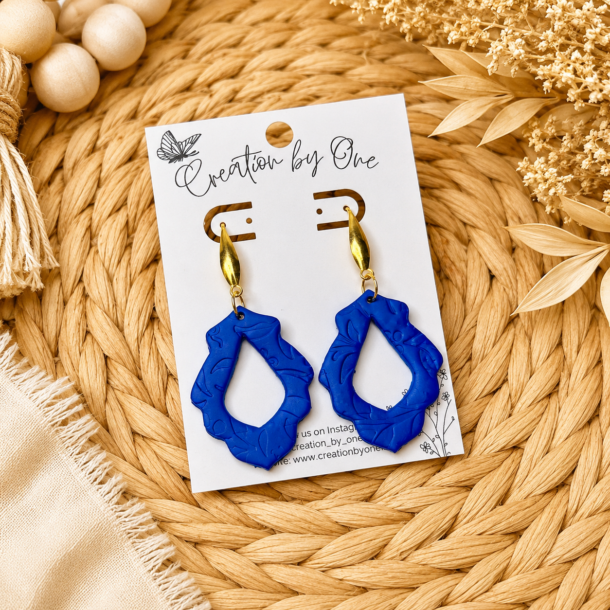 Blue earrings on a 'Creation by One' branded card with a natural background