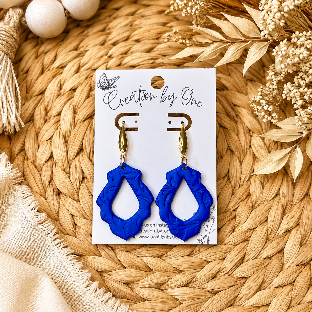 Blue earrings on a white card with 'Creation by One' branding, placed on a woven mat with dried flowers.