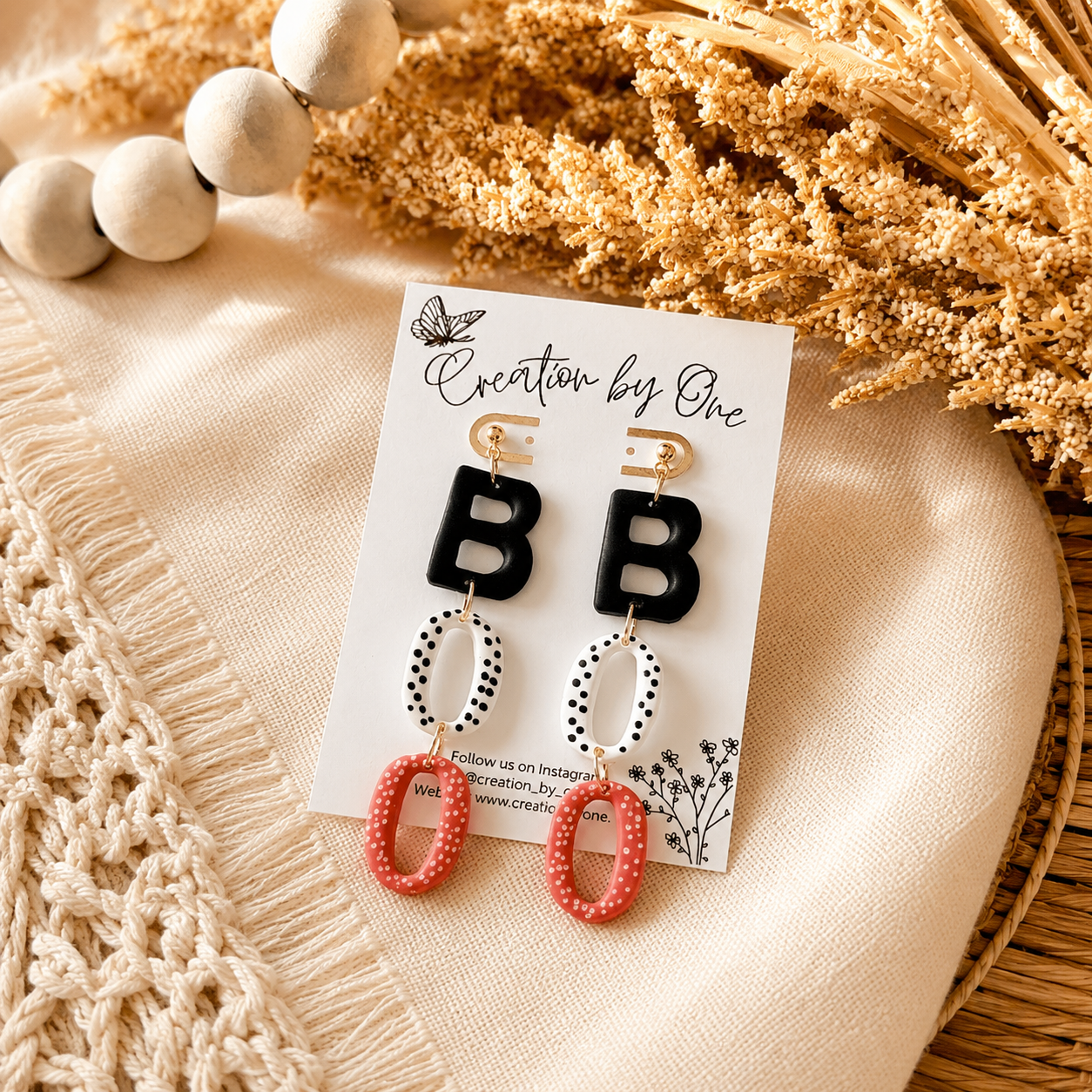 Pair of earrings on a card with 'Creation by One' branding, displayed on a textured surface.