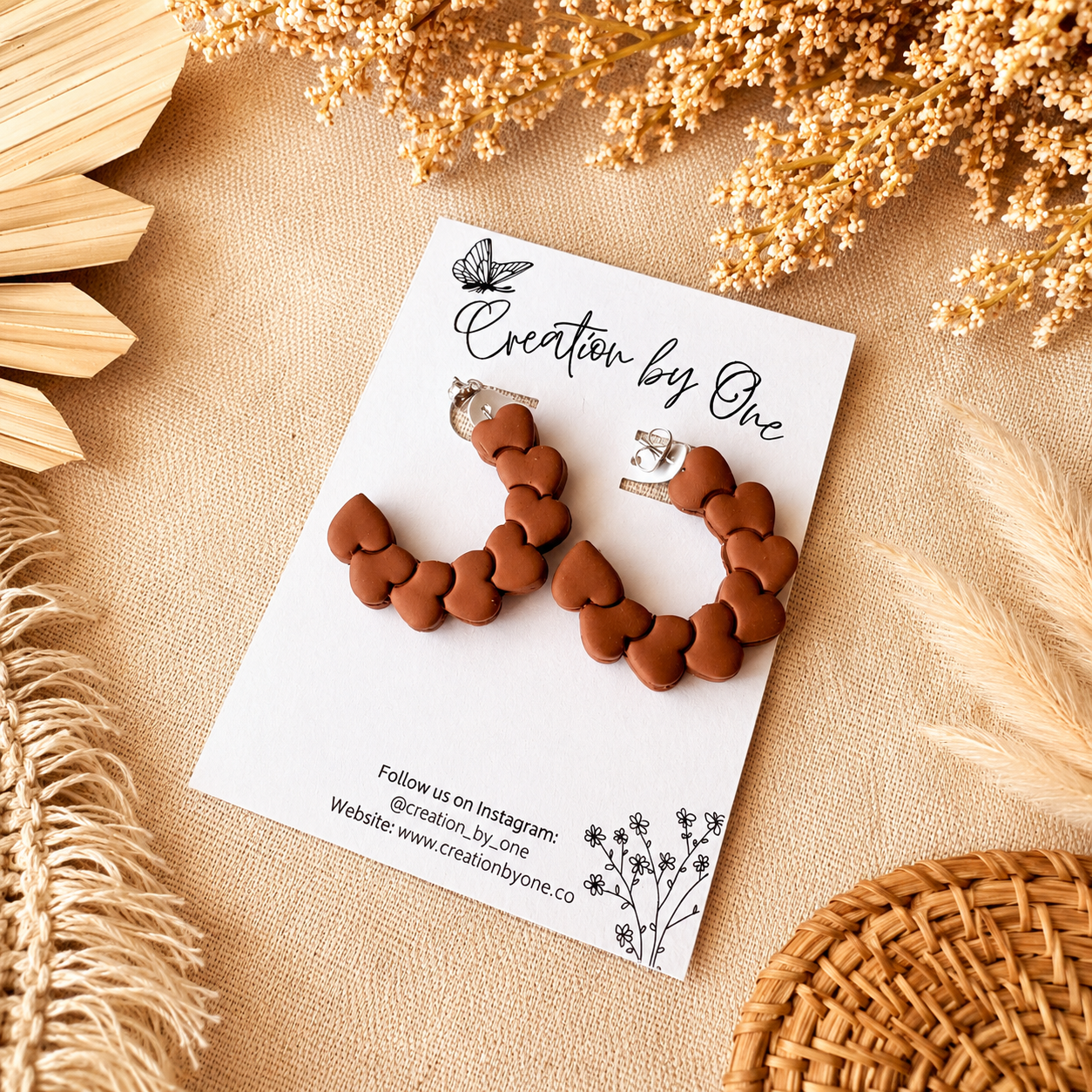 Brown heart-shaped earrings on a 'Creation by One' branded card with dried flowers and natural elements in the background.
