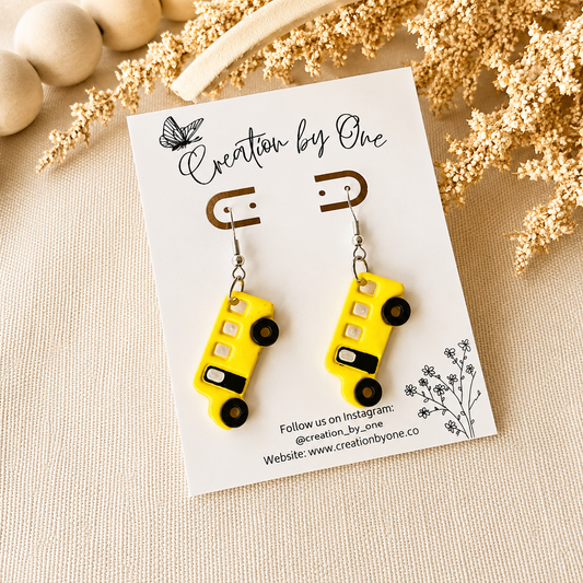 Yellow bus-shaped earrings on a 'Creation by One' branded card with decorative elements.