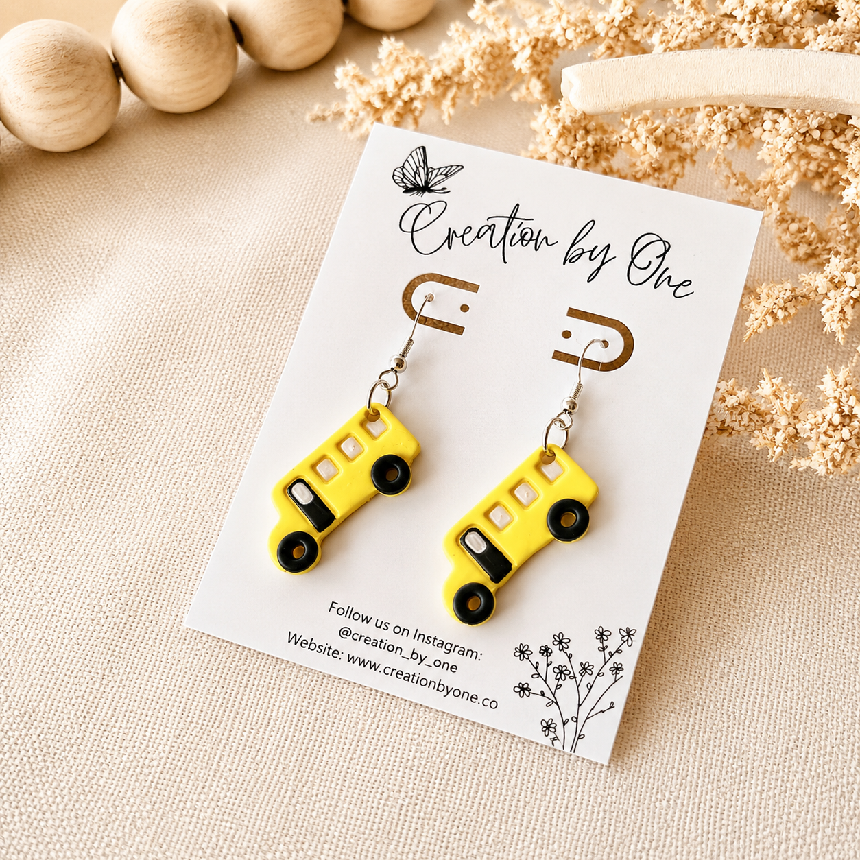 Yellow bus-shaped earrings on a white card with 'Creation by One' branding, placed on a beige surface with wooden beads and dried flowers.