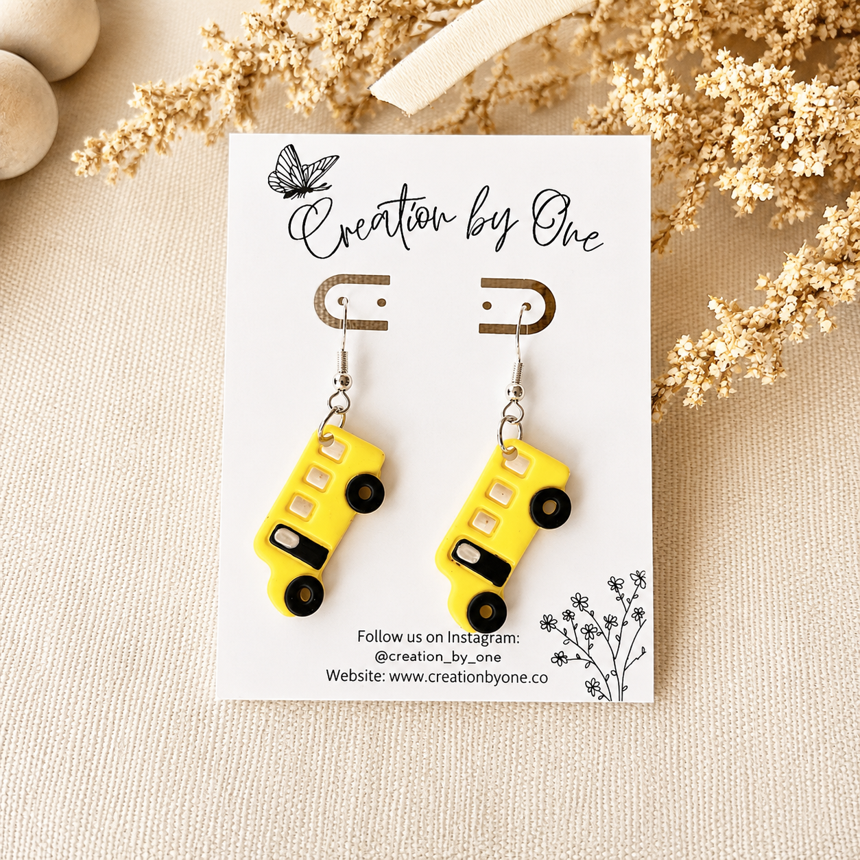 Yellow bus-shaped earrings on a 'Creation by One' branded card with dried flowers in the background.