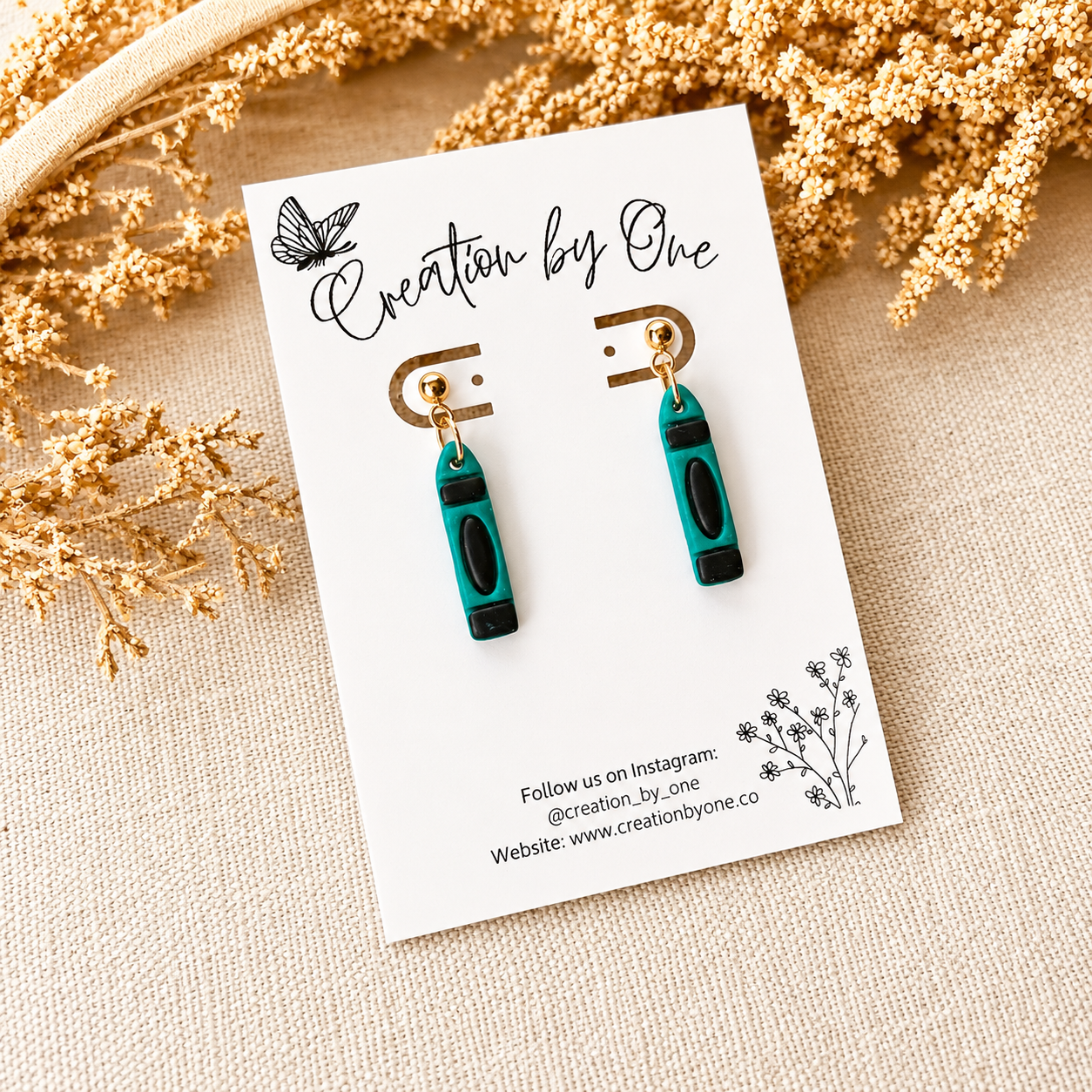 Teal and black earrings on a 'Creation by One' branded card with dried flowers in the background.