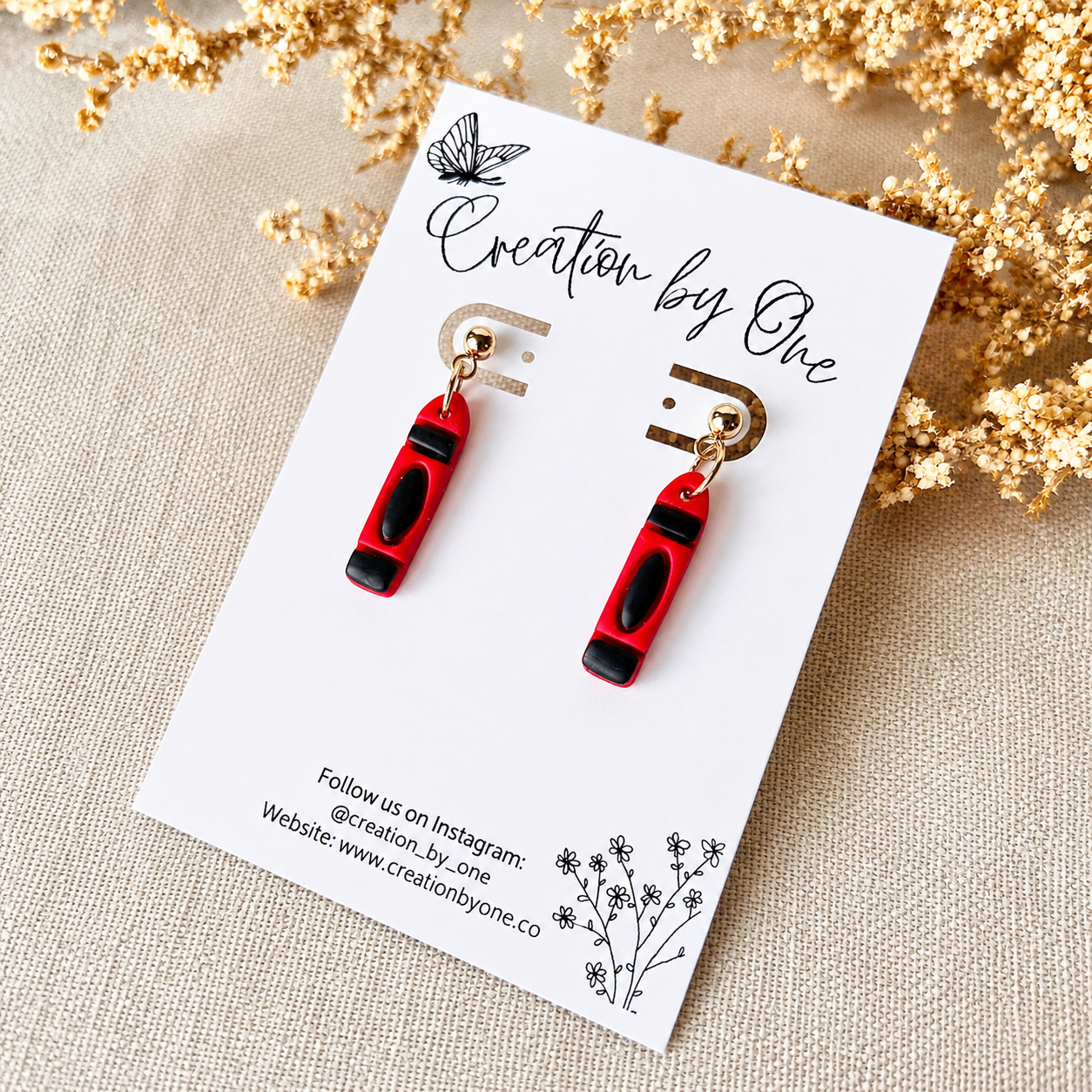 Red and black earrings on a white card with 'Creation by One' branding, set against a beige background with gold flowers.