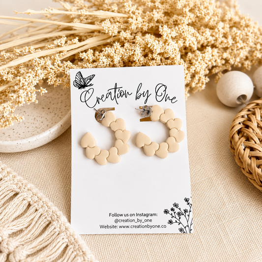 Cream heart hoop earrings on a Creation by One display card with a neutral boho background and dried floral accents.
