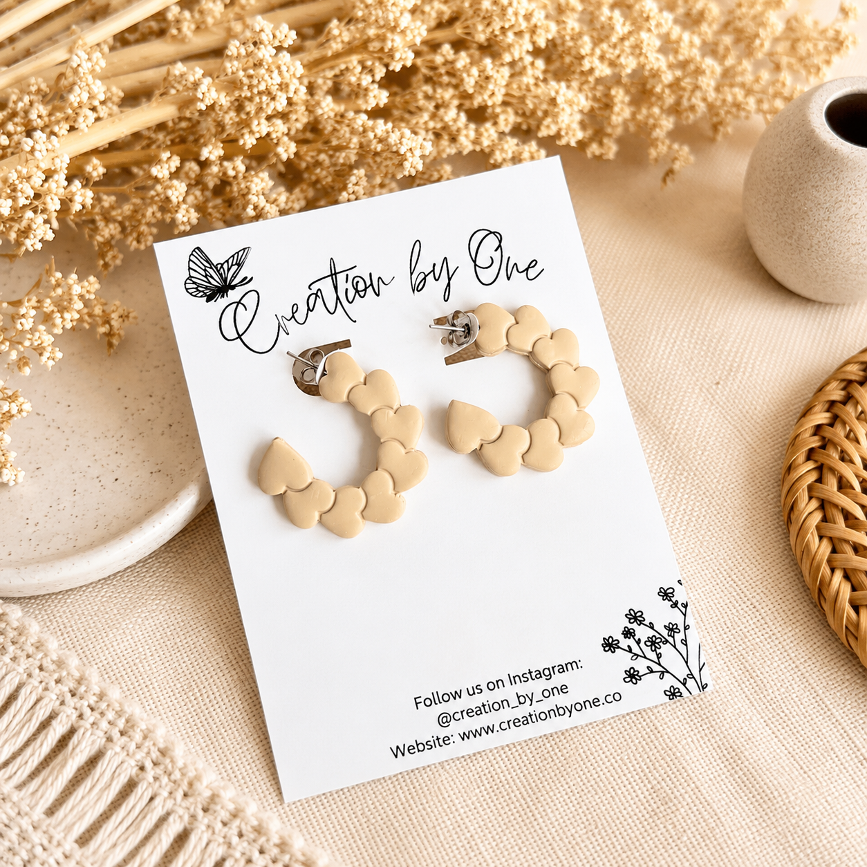Cream heart hoop earrings on a Creation by One display card with a neutral boho background and dried floral accents.