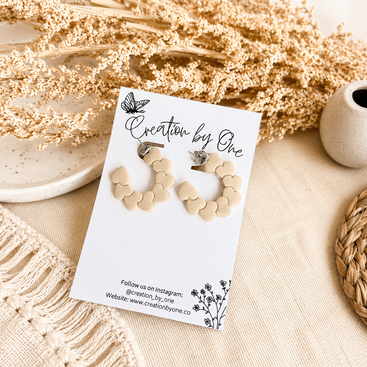 Cream heart hoop earrings on a Creation by One display card with a neutral boho background and dried floral accents.